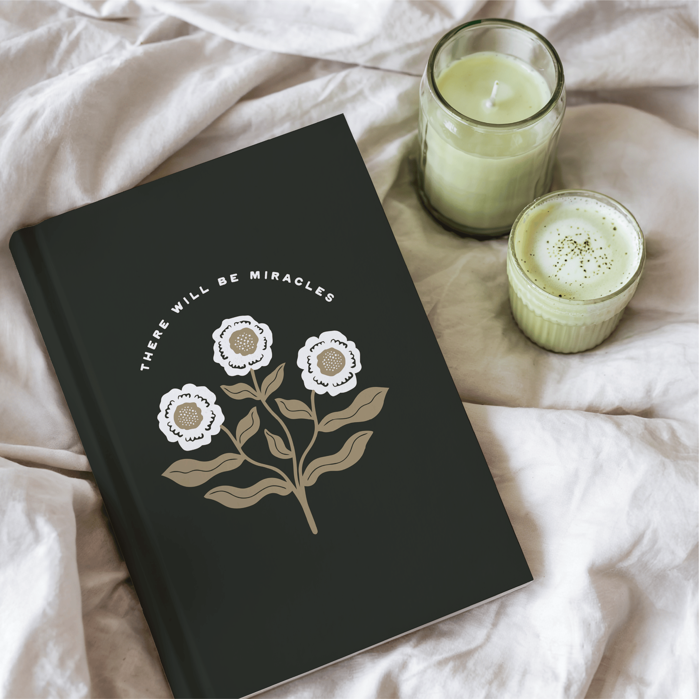 Black notebook with floral design and text, accompanied by two green candles on a light fabric background.