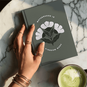 Hand holding a notebook with a floral design on a marble surface with a cup of green tea.