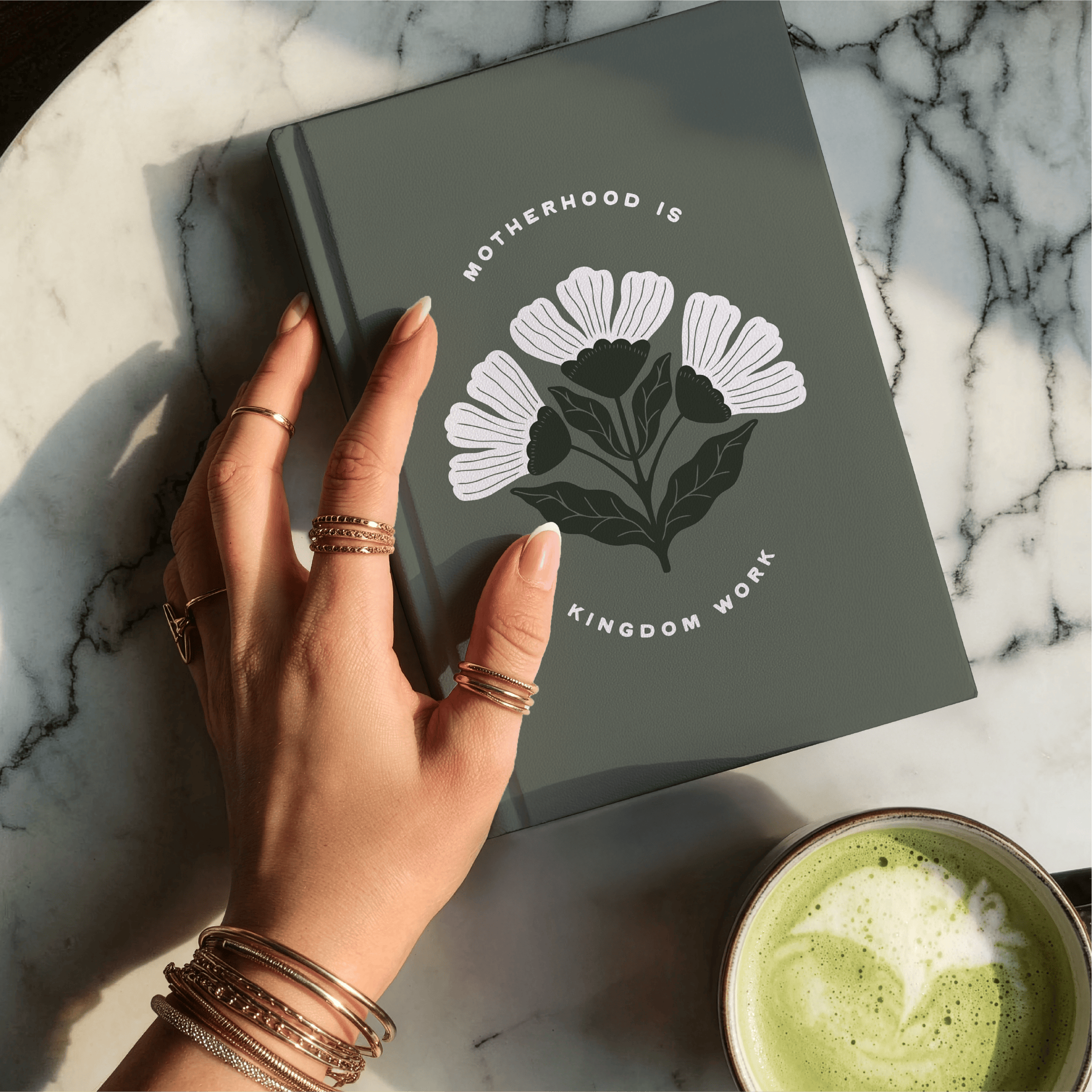 Hand holding a notebook with a floral design on a marble surface with a cup of green tea.