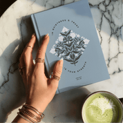 Hand holding a blue notebook with a floral design on a marble surface next to a green beverage.