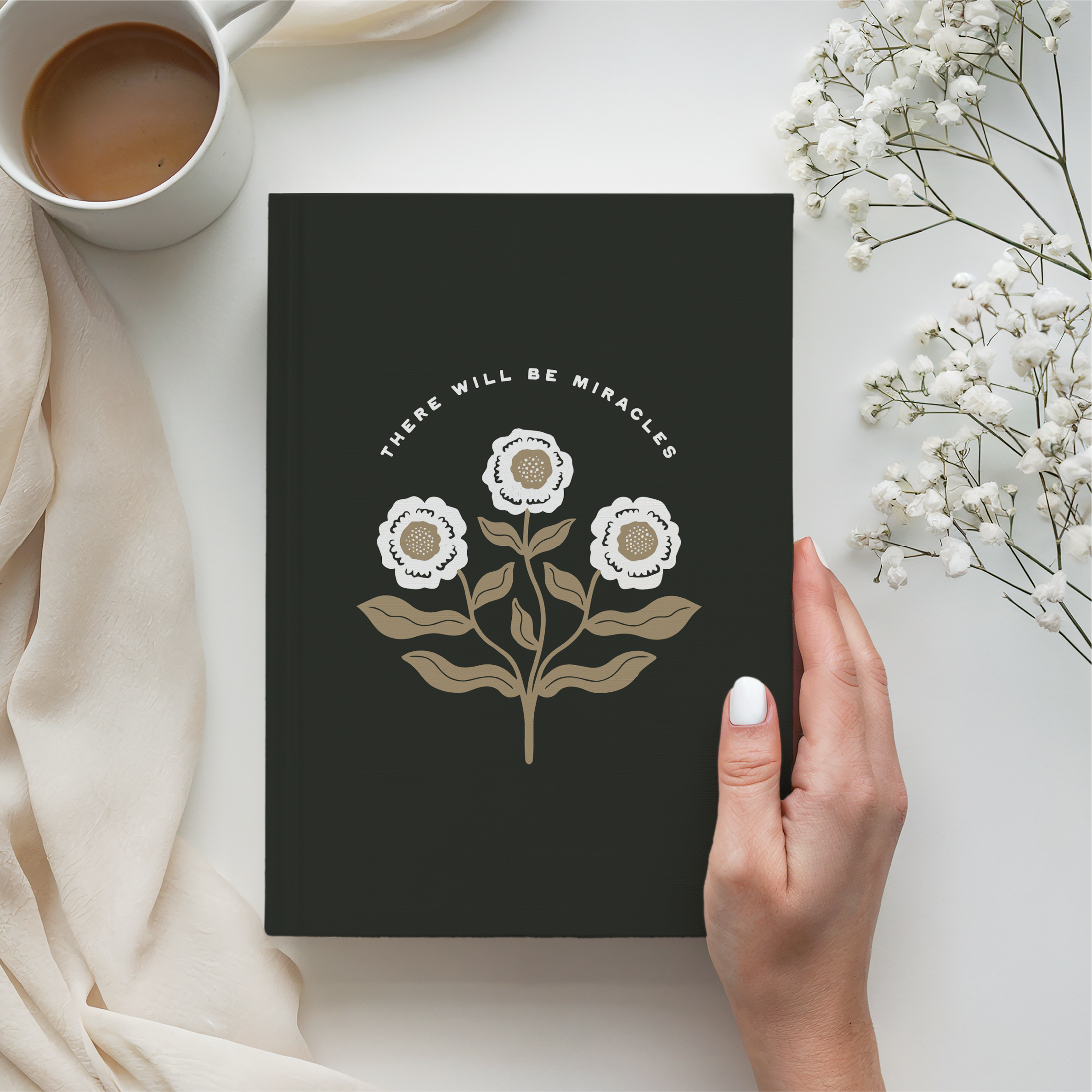 Black notebook with floral design and text, held by a hand, next to a cup of coffee and white flowers on a light surface.