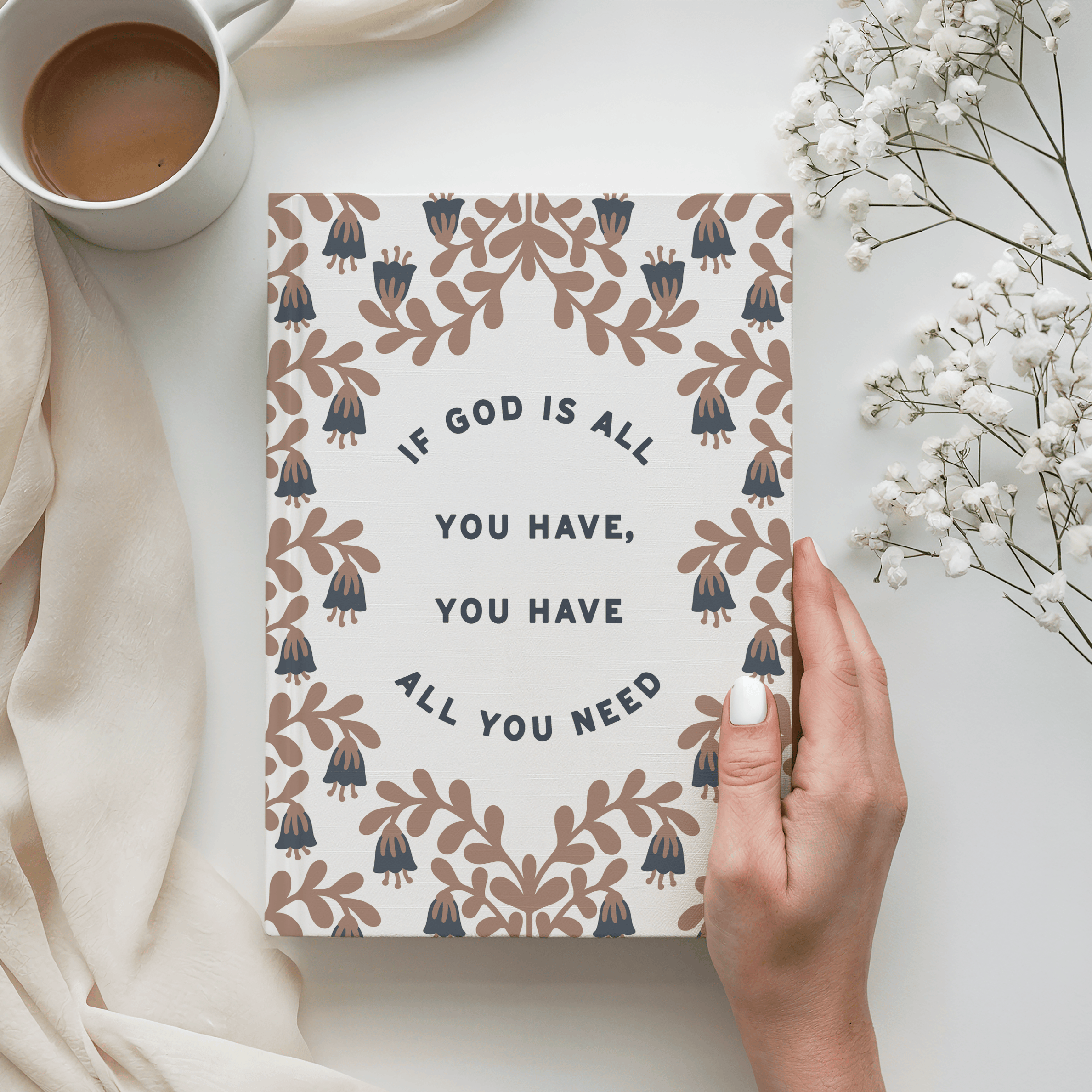 Notebook with motivational quote held by a hand, next to a cup of coffee and flowers on a light surface.