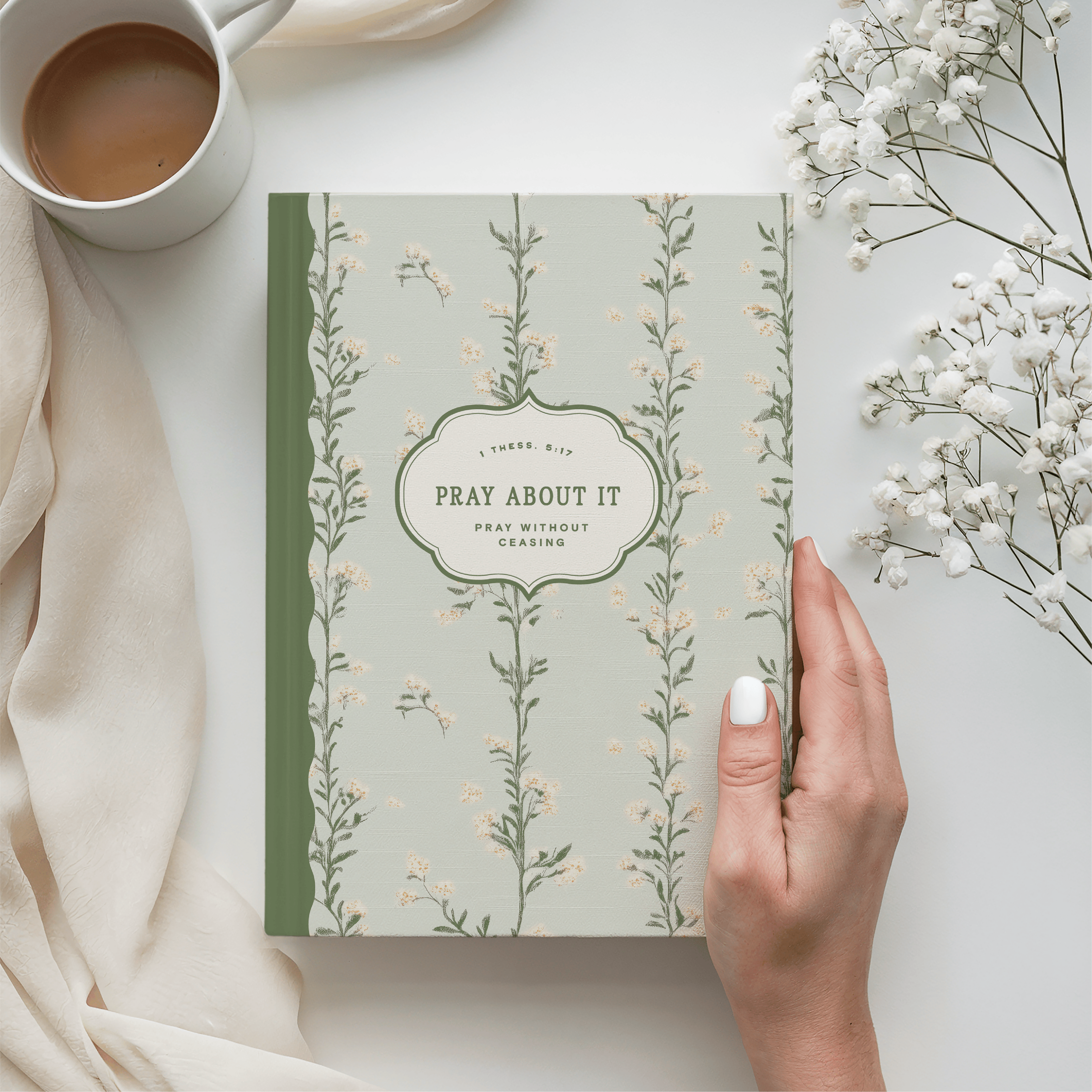 Book titled 'Pray About It' with a hand holding a white flower, cup of coffee, and beige fabric on a light background