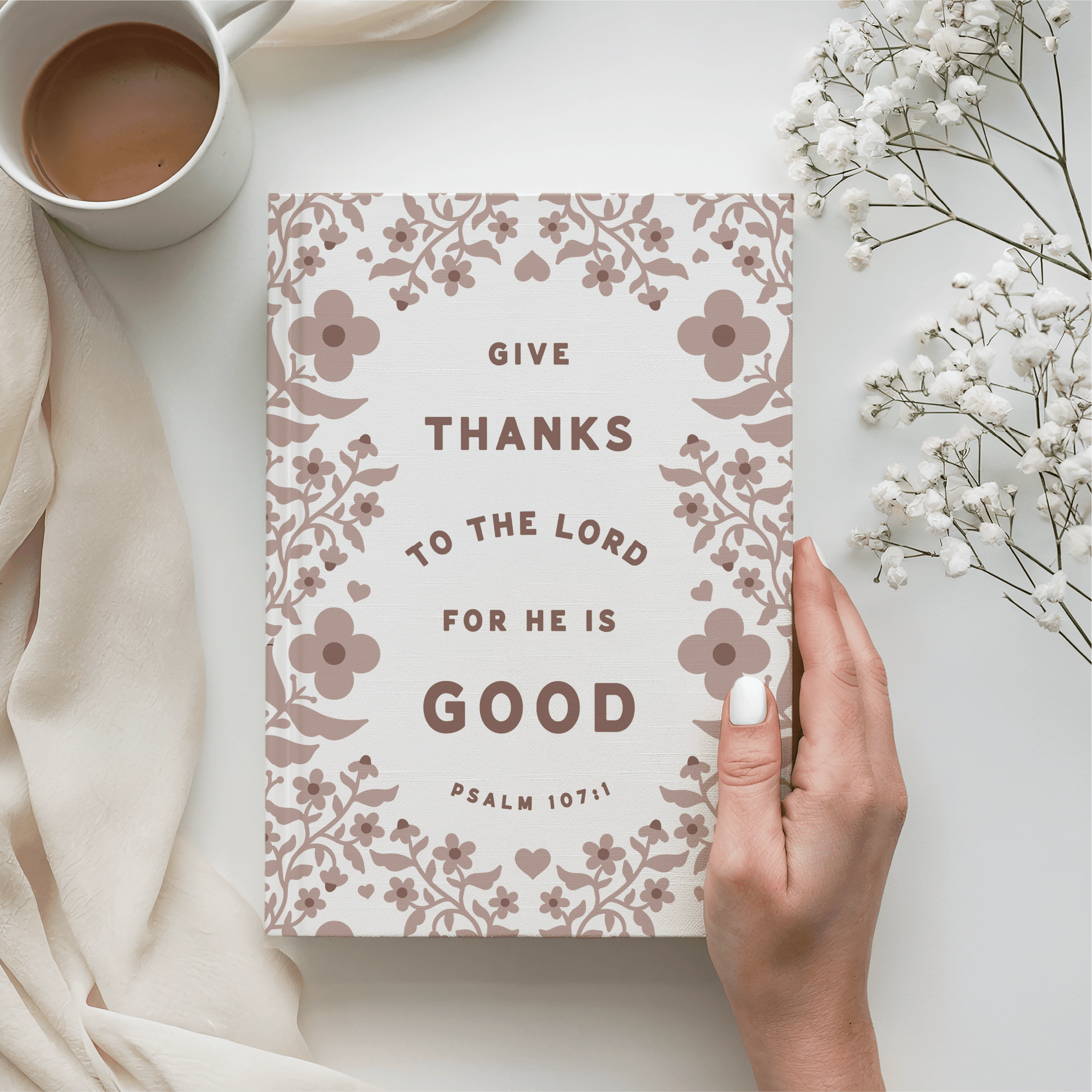 Notebook with 'Give thanks to the Lord for He is Good' text, cup of coffee, and flowers on a light surface