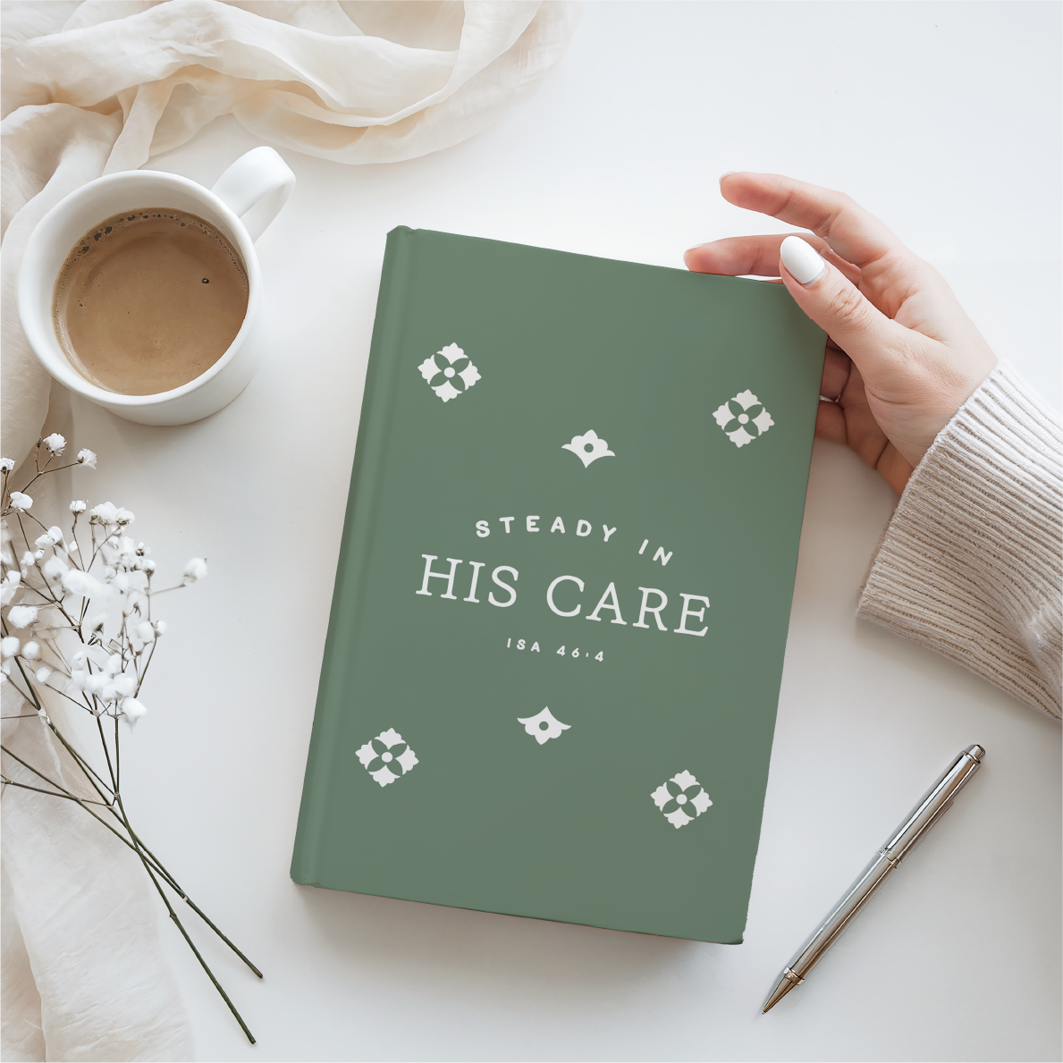Green notebook with 'Steady in His Care' text, held by a hand on a white surface with a cup and pen.