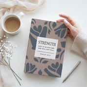 Notebook with 'Strength' quote held by a hand on a white surface with a cup and pen.