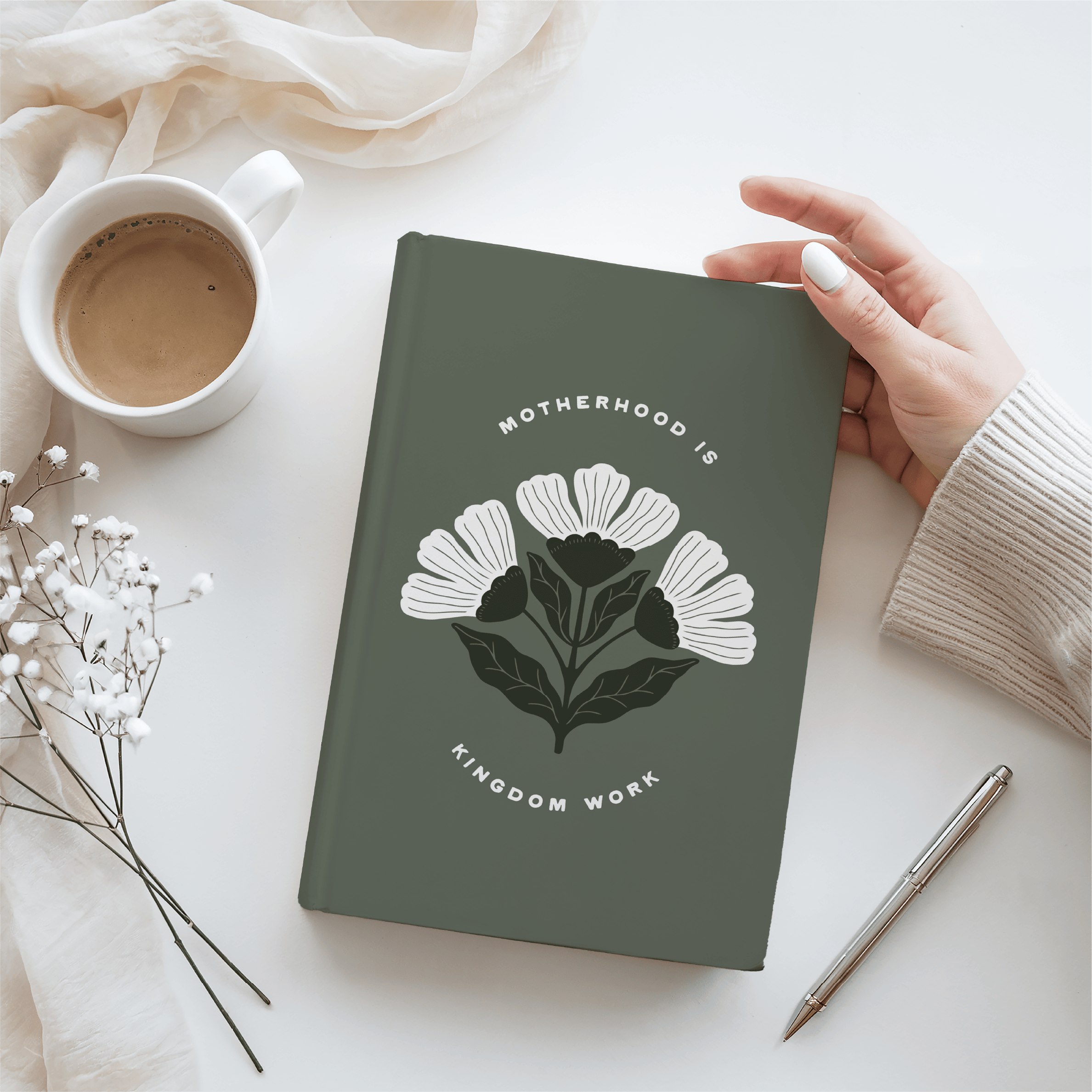 Green notebook with floral design and text, held by a hand on a light surface with a cup and pen.