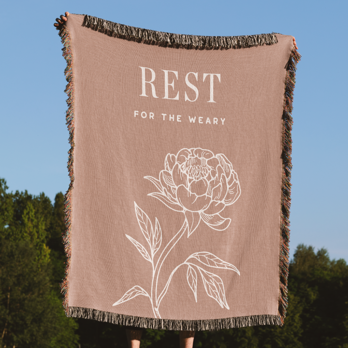 Beige blanket with floral design and 'Rest for the Weary' text against a blue sky and green trees background