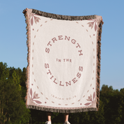 Blanket with 'Strength in the Stillness' text held up against a blue sky and trees