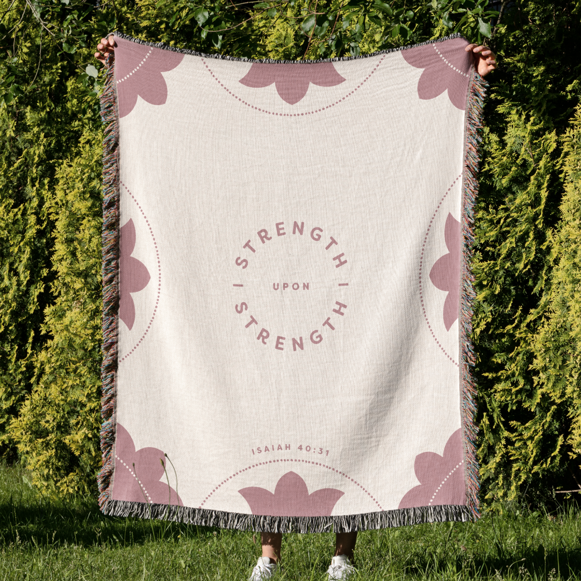 Person holding a blanket with 'Strength is Strength Upon Strength' text against a green outdoor background
