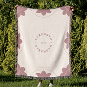 Person holding a blanket with 'Strength is Strength Upon Strength' text against a green outdoor background