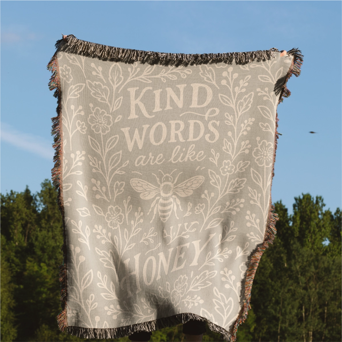 Neutral-toned woven blanket with the phrase “Kind words are like honey,” floral accents, and a honey bee; made of 100% cotton with fringe, perfect Christian gift.