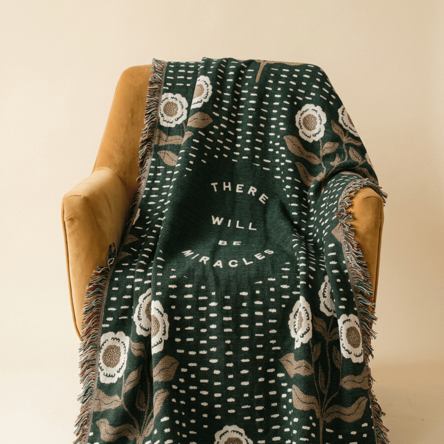 A dark green woven throw blanket with white floral and dotted patterns, draped over a mustard yellow armchair against a warm beige background. The center of the blanket features the text 'THERE WILL BE MIRACLES' in white.