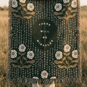 A woven throw blanket with fringe, featuring a dark forest green background with white dotted vertical lines, white flowers, and tan botanical stems. In the center is a dark green oval with the text 'THERE WILL BE MIRACLES' in white.