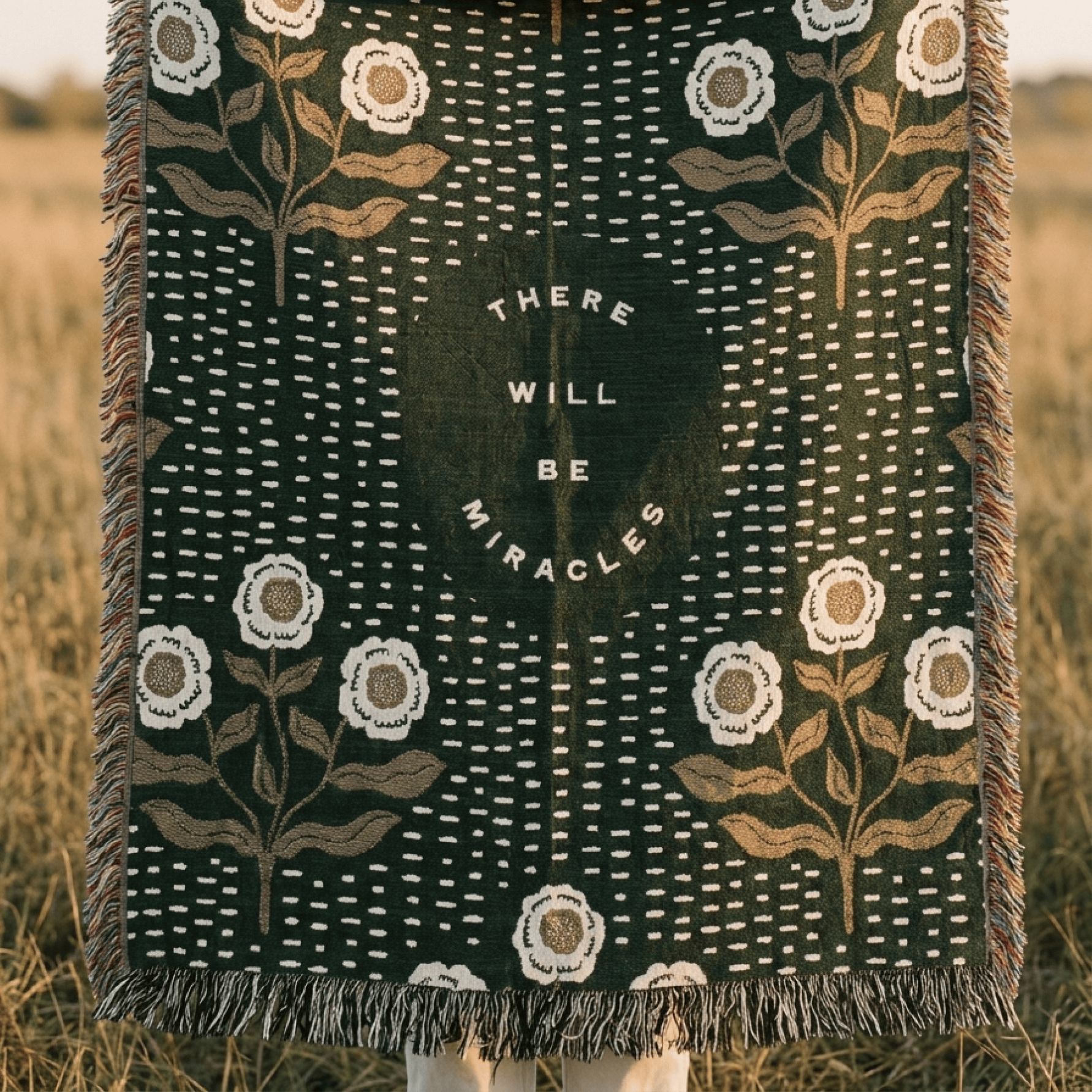 A woven throw blanket with fringe, featuring a dark forest green background with white dotted vertical lines, white flowers, and tan botanical stems. In the center is a dark green oval with the text 'THERE WILL BE MIRACLES' in white.