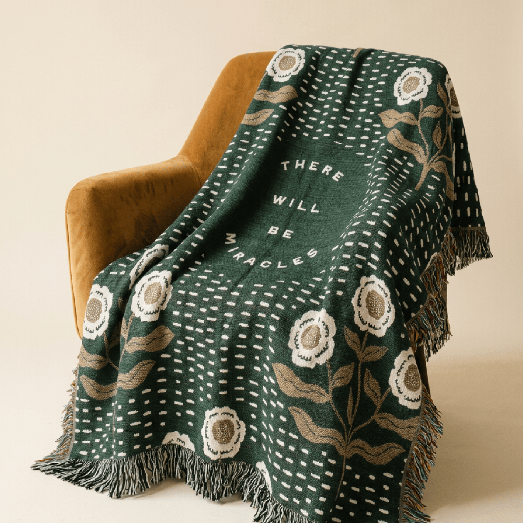 A dark green woven throw blanket with white dotted patterns, beige floral motifs, and the words 'THERE WILL BE MIRACLES' draped over a mustard yellow armchair against a warm beige background.
