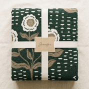 A folded dark green woven blanket or throw with a floral and dotted pattern, wrapped in a white ribbon with a paper tag reading 'Jennifer', photographed from above on a light fabric background.
