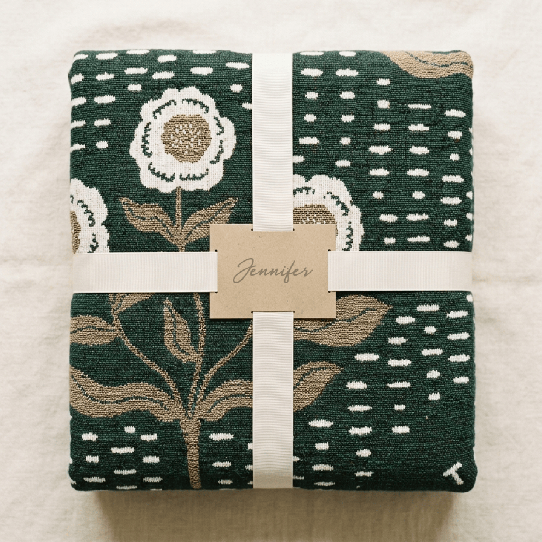 A folded dark green woven blanket or throw with a floral and dotted pattern, wrapped in a white ribbon with a paper tag reading 'Jennifer', photographed from above on a light fabric background.