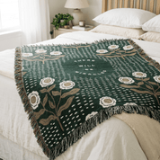 A dark green woven throw blanket with white and beige floral motifs and fringe, draped over a bed in a softly lit bedroom.