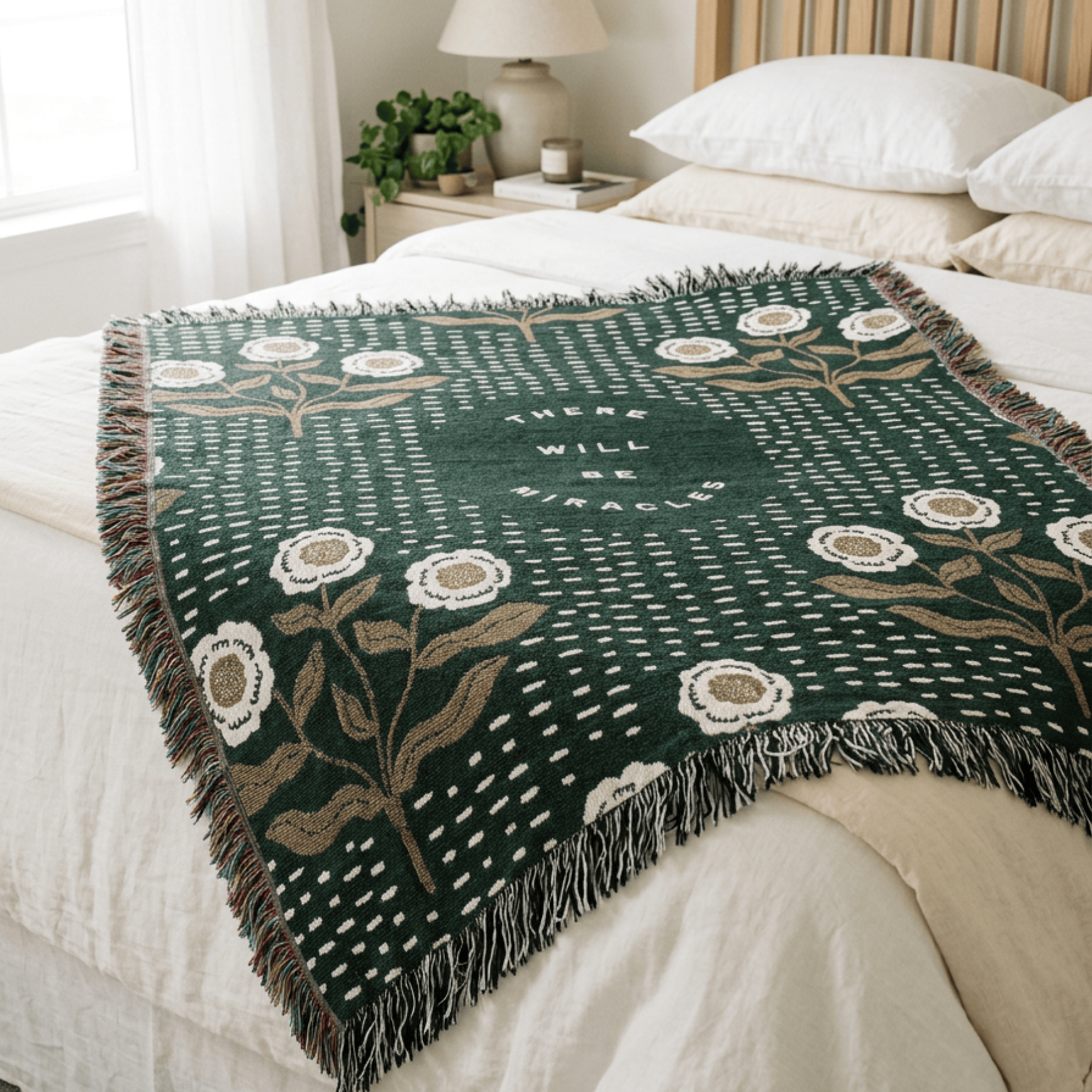 A dark green woven throw blanket with white and beige floral motifs and fringe, draped over a bed in a softly lit bedroom.