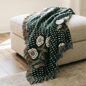 A dark green woven throw blanket with cream and beige floral motifs and fringe, draped over a light beige upholstered ottoman in a softly lit room.