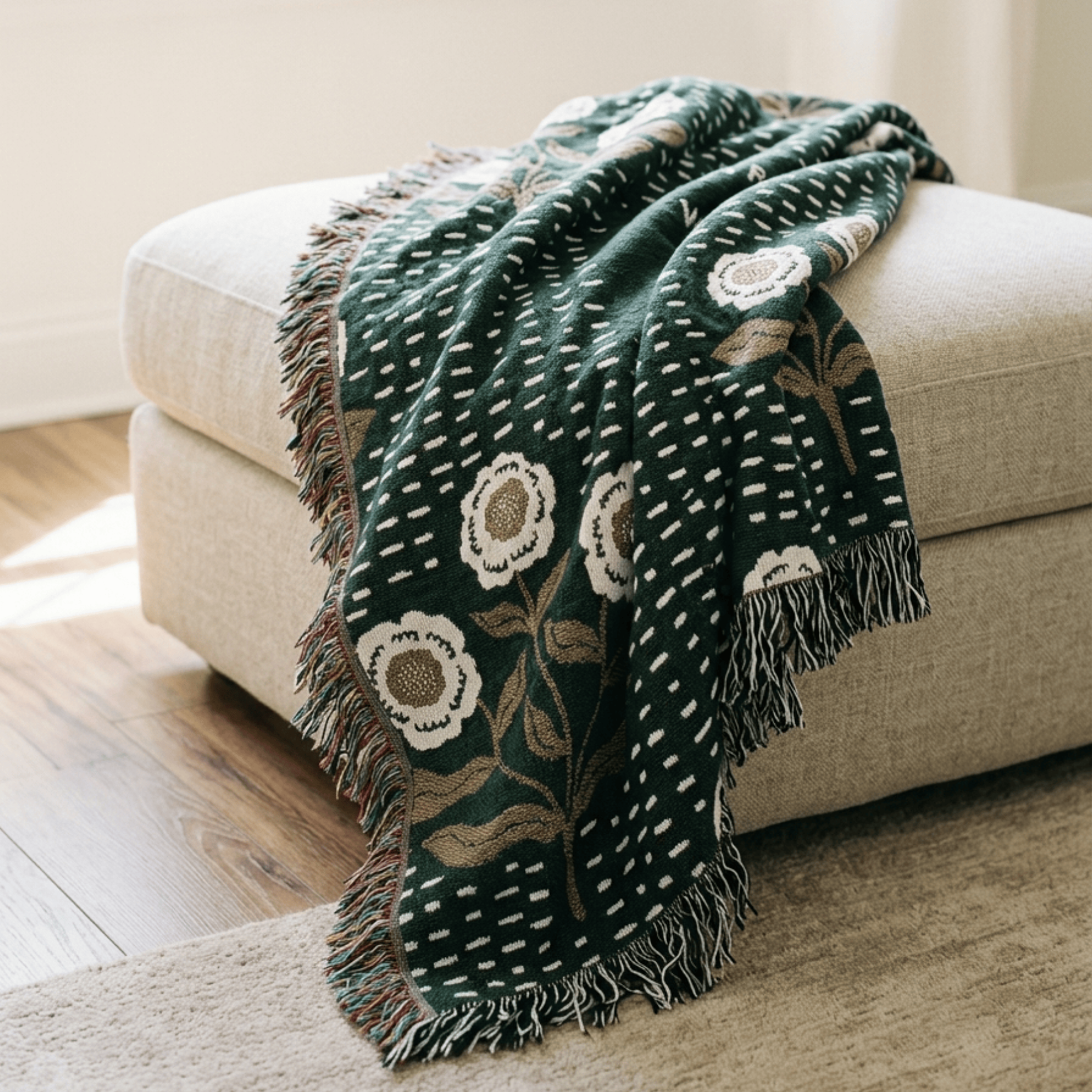 A dark green woven throw blanket with cream and beige floral motifs and fringe, draped over a light beige upholstered ottoman in a softly lit room.