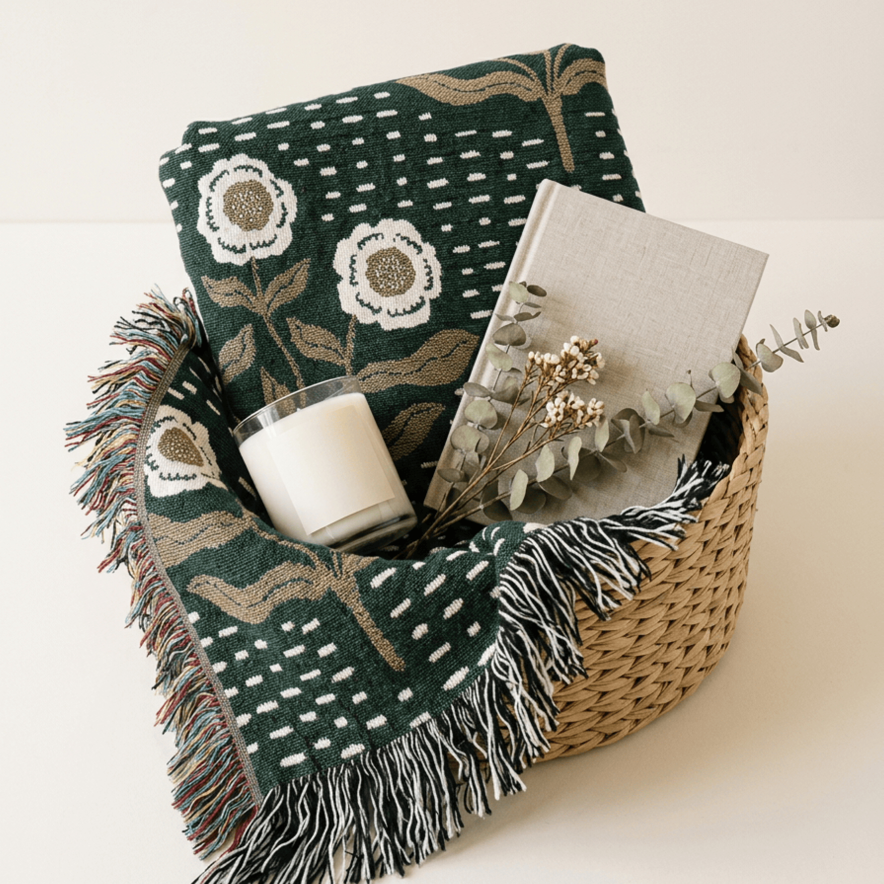 A dark green woven throw blanket with white and beige floral and geometric patterns, styled in a natural woven basket with a candle, notebook, and dried eucalyptus stems on a light beige background.