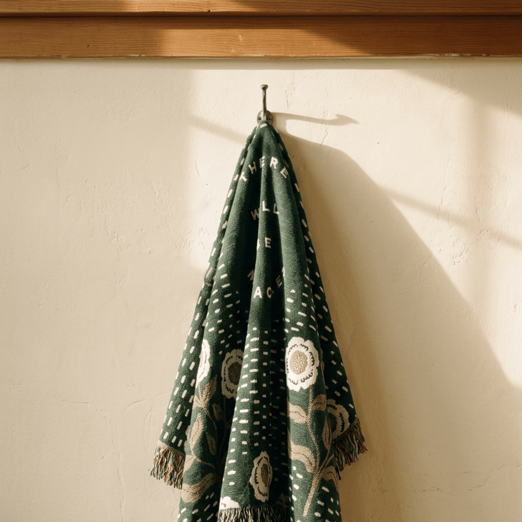 A dark green woven throw blanket with white and beige floral patterns and fringe, hanging from a hook on a textured cream wall.