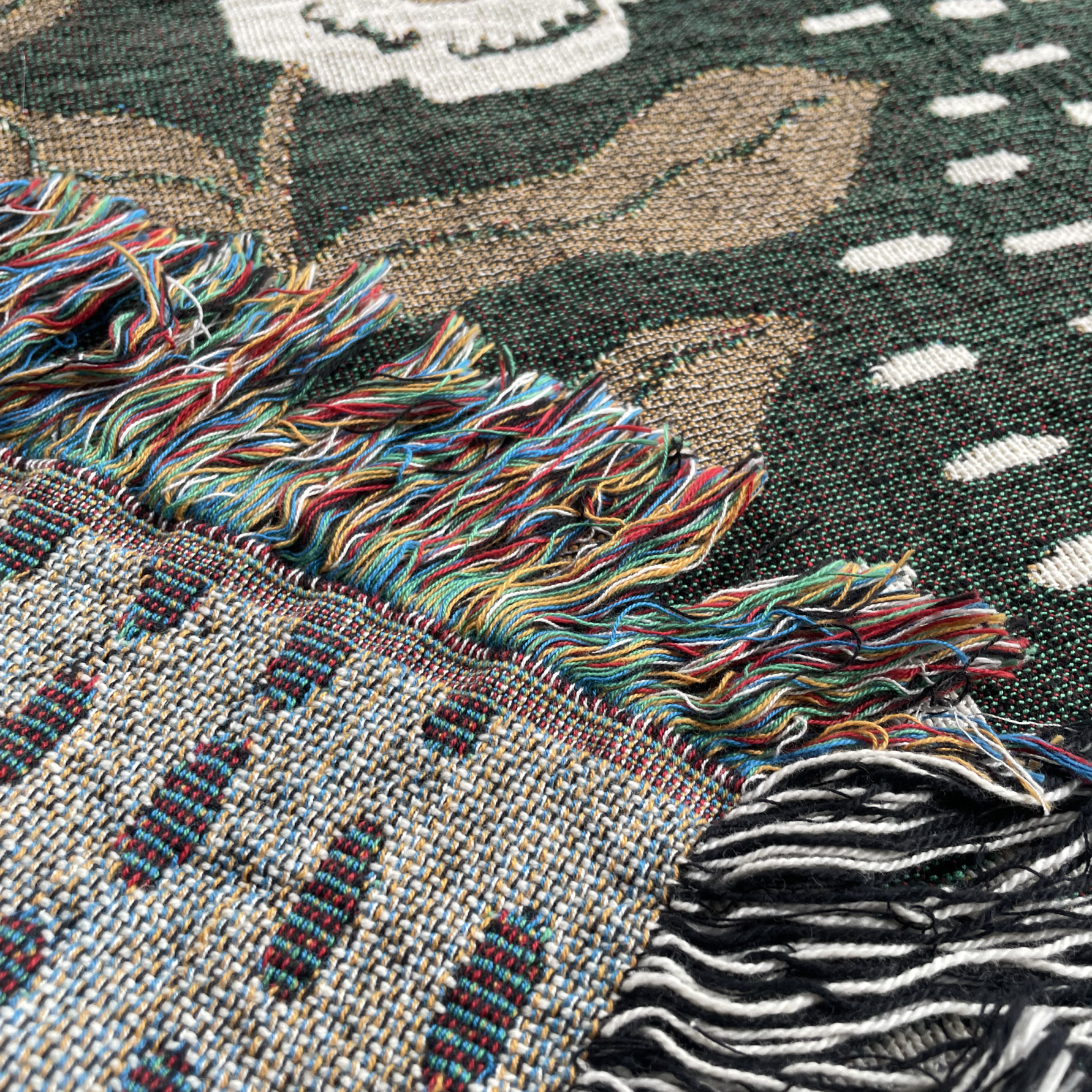 Close-up of a woven textile with multicolored fringe, showing a dark green patterned section and a beige-and-gray patterned section with black-and-white fringe.