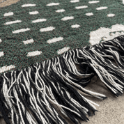 Close-up of a woven textile with a dark green base, irregular white dotted pattern, and black-and-white fringe tassels on a neutral surface.