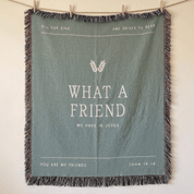 A square sage green woven throw blanket with fringe, hanging from a wooden clothesline with wooden clips against a warm off-white wall. The blanket features white printed text and a wheat graphic.