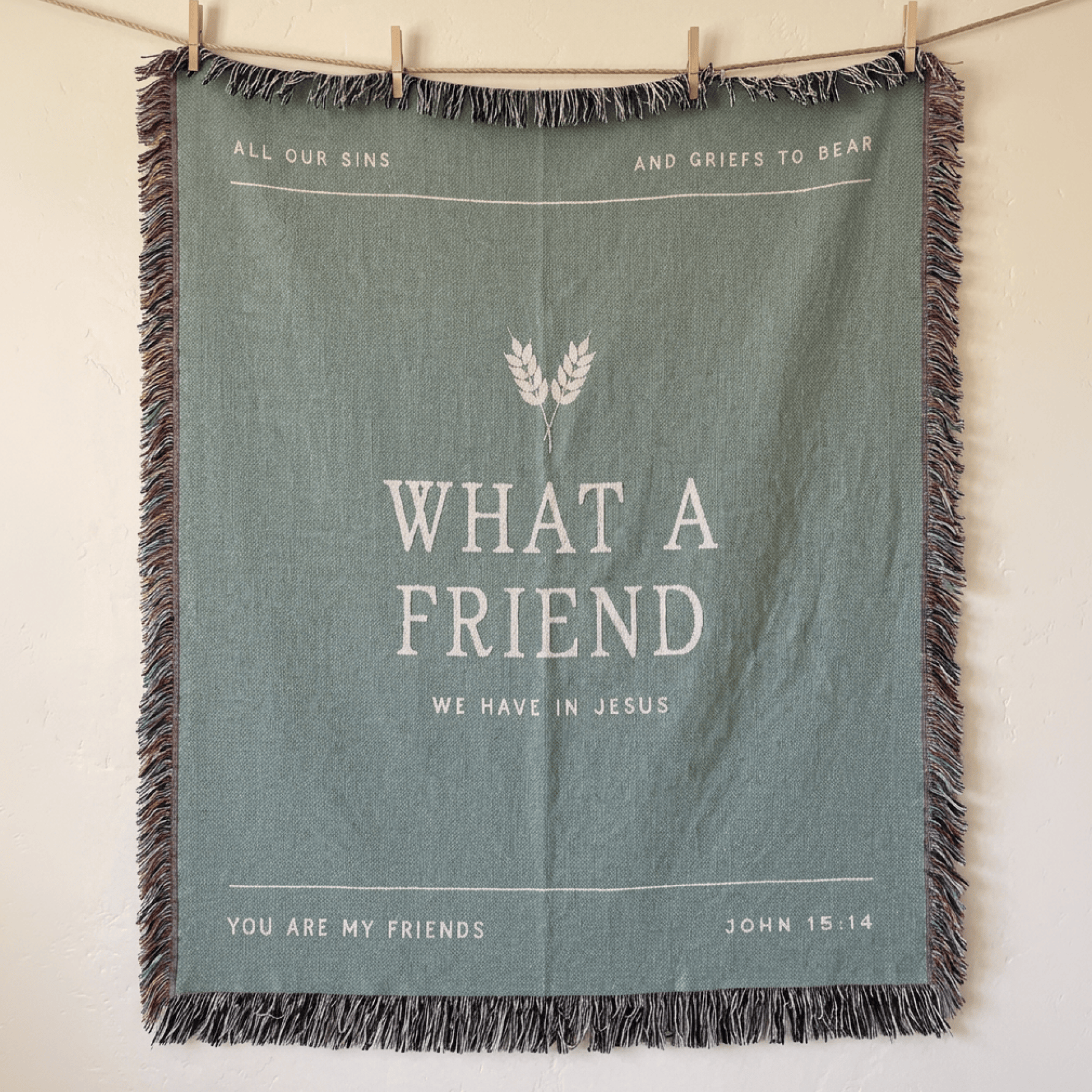 A square sage green woven throw blanket with fringe, hanging from a wooden clothesline with wooden clips against a warm off-white wall. The blanket features white printed text and a wheat graphic.
