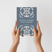 Book titled 'Be Still & Know That I Am God' held by hands against a light gray background
