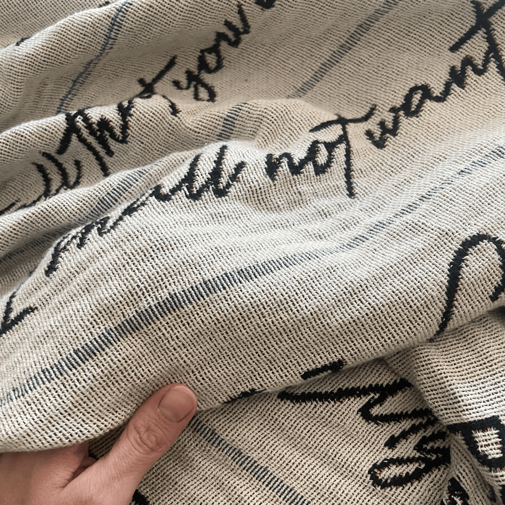 A close-up of a woven textile with a cream base, black cursive script text, and thin gray stripes, held by a hand to show texture and drape.