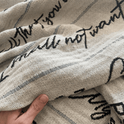 A close-up of a woven textile with a cream base, black cursive script text, and thin gray stripes, held by a hand to show texture and drape.