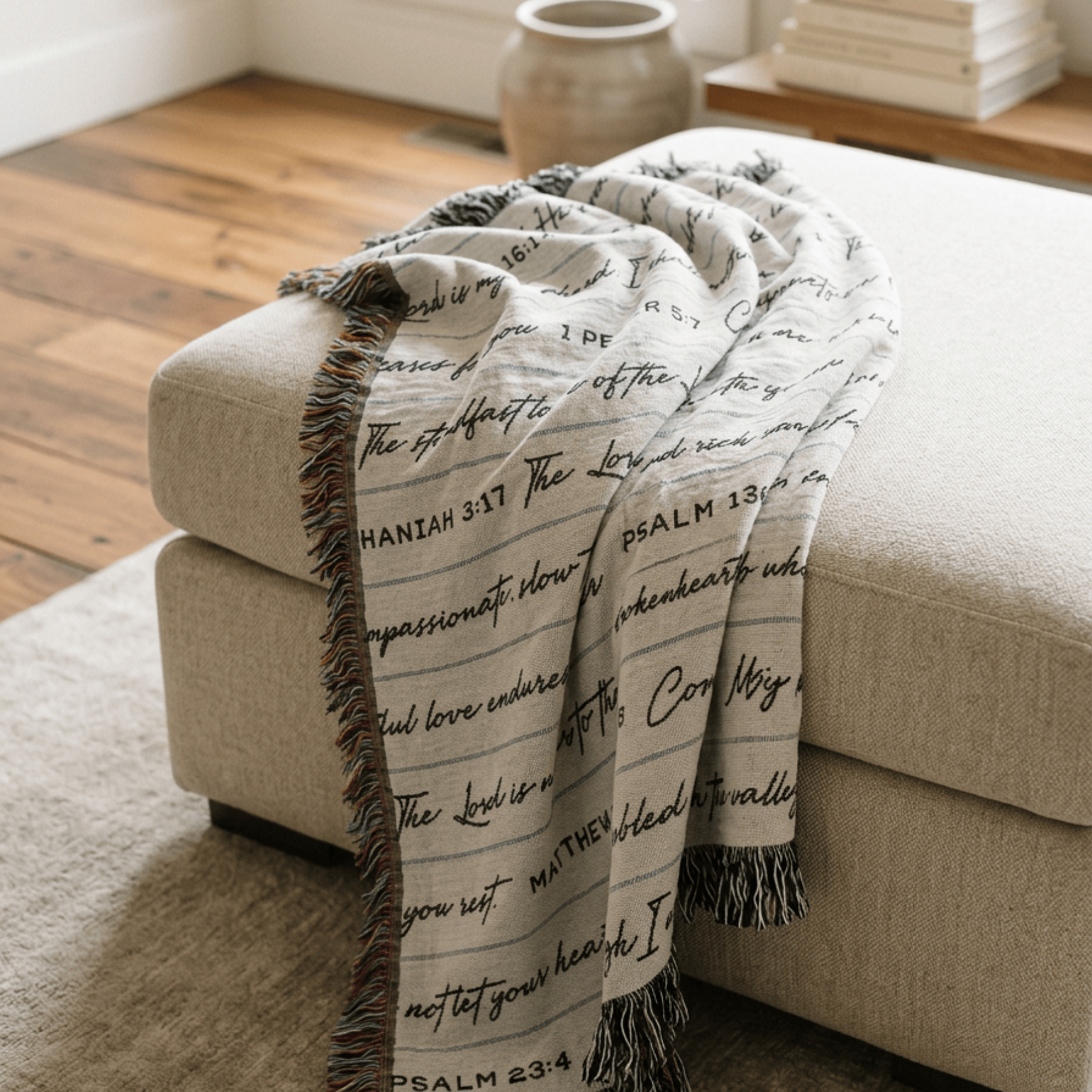 A cream-colored throw blanket with black cursive Bible verse text and fringed edges, draped over a beige fabric ottoman in a cozy room setting with wooden floors.