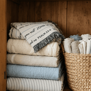 A stack of folded woven textiles in neutral and soft blue tones sits inside a wooden cabinet next to a woven basket holding more folded linens.