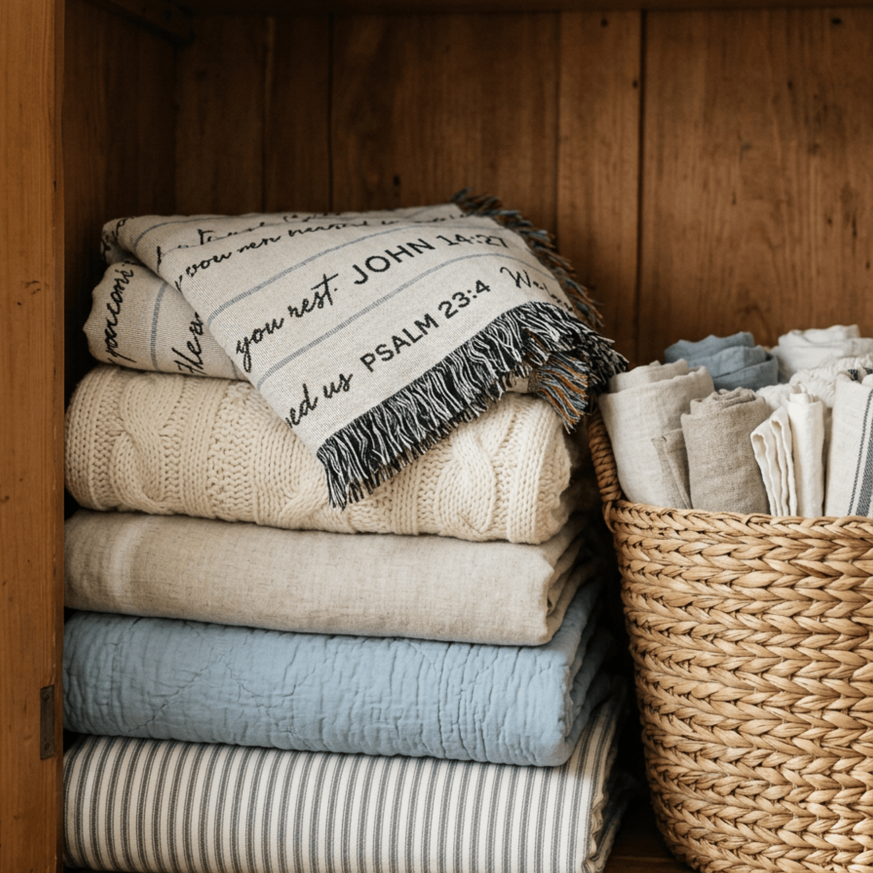 A stack of folded woven textiles in neutral and soft blue tones sits inside a wooden cabinet next to a woven basket holding more folded linens.